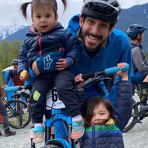 Jared Sartee smiling with kids hanging of mountain bike handlebars
